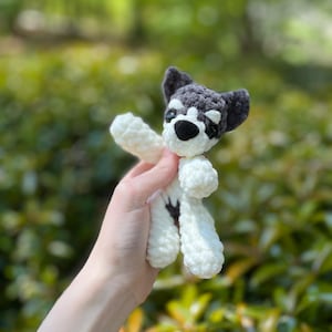 May include: A handmade crochet dog toy in white and gray. The dog has a black nose and eyes, with gray ears and a gray patch over one eye. The toy is held against a blurred green background.