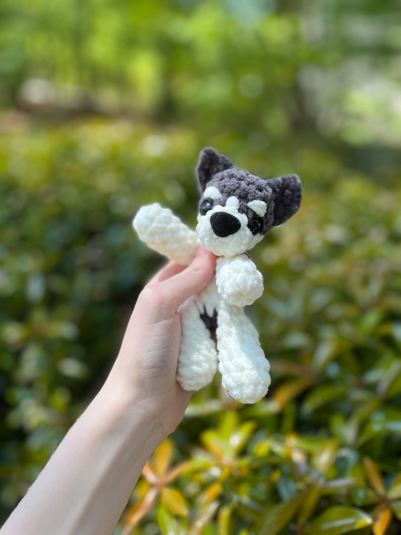 May include: A handmade crochet dog toy in white and gray. The dog has a black nose and eyes, with gray ears and a gray patch over one eye. The toy is held against a blurred green background.