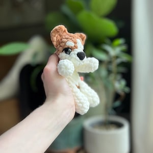 May include: A crocheted dog toy in white, brown, and black. The small stuffed animal has a white body, brown head and ears, and black nose and eyes. The toy is held up in front of a blurred background of plants.