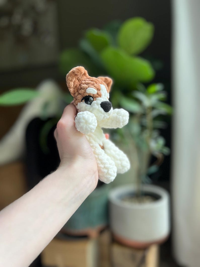 May include: A crocheted dog toy in white, brown, and black. The small stuffed animal has a white body, brown head and ears, and black nose and eyes. The toy is held up in front of a blurred background of plants.