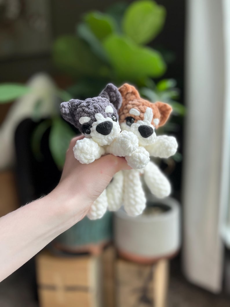 May include: Two crocheted husky dog plushies. One is gray and white, the other is brown and white. Both have black eyes and noses. The plushies are held in a hand, with a blurred background of plants and a window.