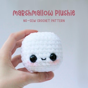 以下が含まれることがあります： 黒い目とピンクの頬をした、白の毛糸で編まれたマシュマロのぬいぐるみ。ぬいぐるみは手で持たれています。ぬいぐるみの上に「marshmallow plushie」と「no-sew crochet pattern」の文字があります。