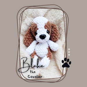 Puede incluir: Un peluche de perro Cavalier King Charles Spaniel hecho a mano con ganchillo. El peluche es marrón, blanco y negro, con el nombre "Blake the Cavalier" escrito en cursiva. El peluche está sobre una manta crema y esponjosa.