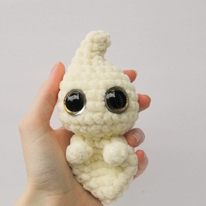 Puede incluir: Un juguete de peluche fantasma blanco tejido a crochet con grandes ojos negros y detalles dorados. El fantasma est&aacute; siendo sostenido en la mano de una persona.