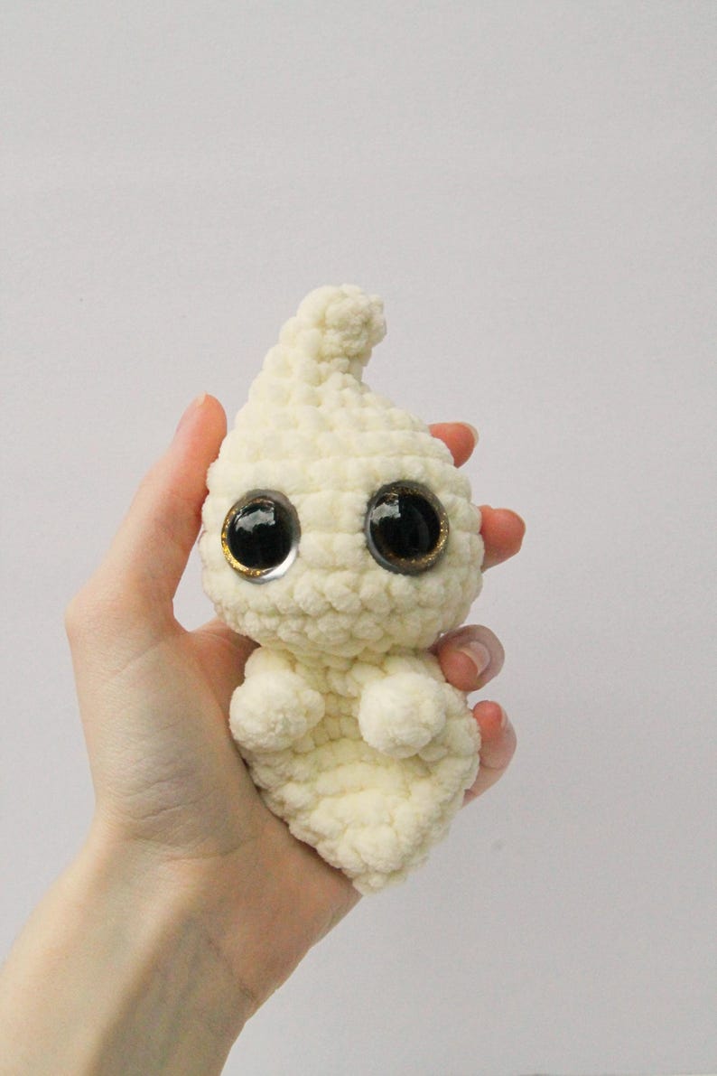 Puede incluir: Un juguete de peluche fantasma blanco tejido a crochet con grandes ojos negros y detalles dorados. El fantasma est&aacute; siendo sostenido en la mano de una persona.