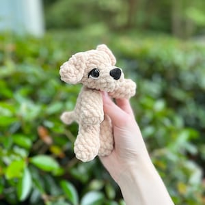 以下が含まれることがあります： 柔らかい質感の糸で編まれた、ベージュ色の小さなかぎ針編みの犬のぬいぐるみ。垂れ耳、小さな黒い鼻、暗い色の丸いプラスチック製の目が特徴です。犬は4本の足で立っているシンプルな姿勢で、後ろには短い尻尾が見えます。