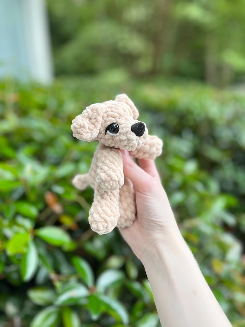 以下が含まれることがあります： 柔らかい質感の糸で編まれた、ベージュ色の小さなかぎ針編みの犬のぬいぐるみ。垂れ耳、小さな黒い鼻、暗い色の丸いプラスチック製の目が特徴です。犬は4本の足で立っているシンプルな姿勢で、後ろには短い尻尾が見えます。