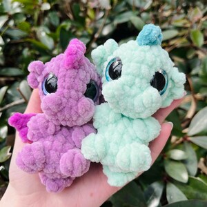 May include: Two handmade crochet dragon plushies. One is lavender with pink accents, the other is mint green with a blue accent. Both have large, shiny black eyes. The dragons are held in a hand against a backdrop of green foliage.