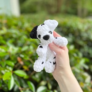 以下が含まれることがあります： ダルメシアン犬の形をした、手編みの小さなかぎ針編みのぬいぐるみ。このぬいぐるみは、白く柔らかい質感の毛糸で作られており、体全体に黒い斑点があり、片方の耳は黒く、黒い鼻と暗い丸いプラスチック製の目が付いています。