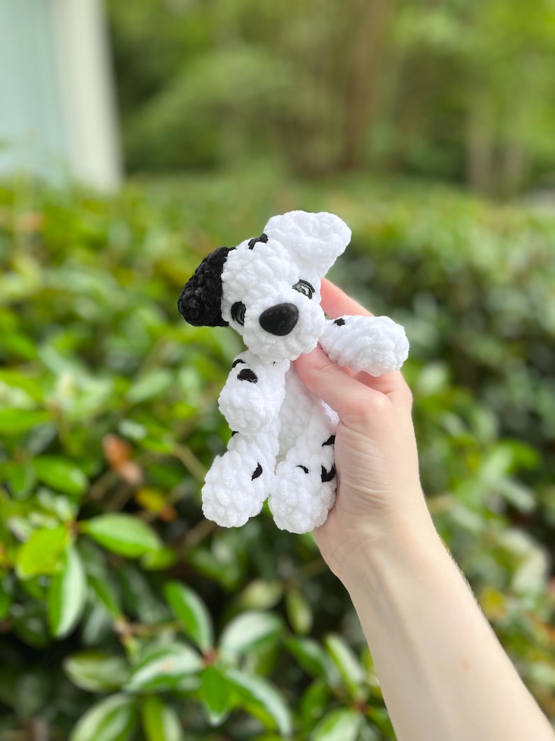 以下が含まれることがあります： ダルメシアン犬の形をした、手編みの小さなかぎ針編みのぬいぐるみ。このぬいぐるみは、白く柔らかい質感の毛糸で作られており、体全体に黒い斑点があり、片方の耳は黒く、黒い鼻と暗い丸いプラスチック製の目が付いています。