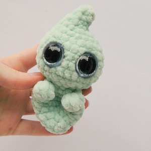 Puede incluir: Un peque&ntilde;o juguete de peluche tejido a crochet con forma de fantasma. El fantasma es de color verde claro con ojos grandes y negros con purpurina azul.