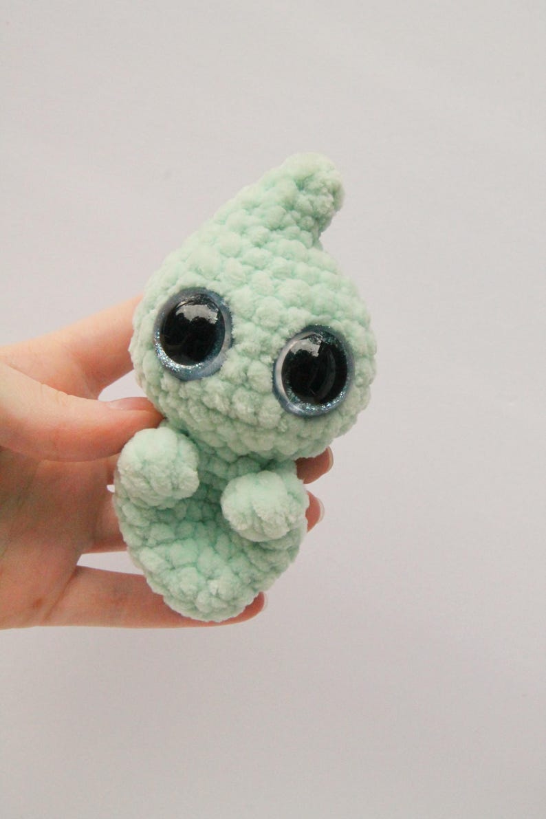 Puede incluir: Un peque&ntilde;o juguete de peluche tejido a crochet con forma de fantasma. El fantasma es de color verde claro con ojos grandes y negros con purpurina azul.