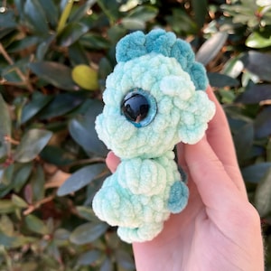 May include: A small, handmade crochet seahorse in shades of light blue and teal. The plush toy features a textured, fluffy body, a detailed head with a dark eye, and a curled tail. The seahorse is held against a backdrop of green foliage.