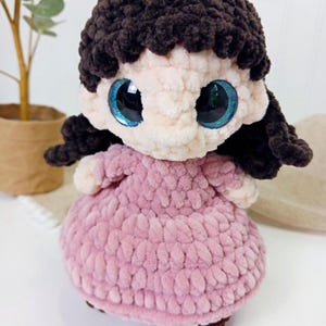 May include: A handmade crochet doll with a light pink dress and dark brown hair. The doll has large, sparkling blue eyes and a soft, textured appearance. The doll is a charming, handcrafted item.