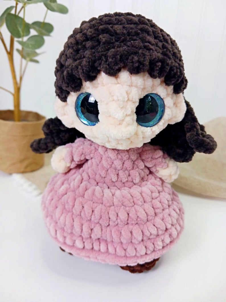 May include: A handmade crochet doll with a light pink dress and dark brown hair. The doll has large, sparkling blue eyes and a soft, textured appearance. The doll is a charming, handcrafted item.