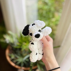 以下が含まれることがあります： ダルメシアン犬の形をした、手編みの小さなかぎ針編みのぬいぐるみ。白い毛糸に黒い斑点があり、黒い耳、黒い鼻、暗い色の目が特徴です。柔らかく厚みのある質感で、丸みを帯びた頭と手足がついています。