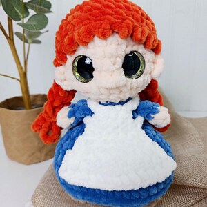 May include: A handmade plush doll with bright orange hair styled in a bun and pigtails. The doll has large, expressive eyes and is wearing a blue dress with a white apron. The doll is made of soft, textured yarn.