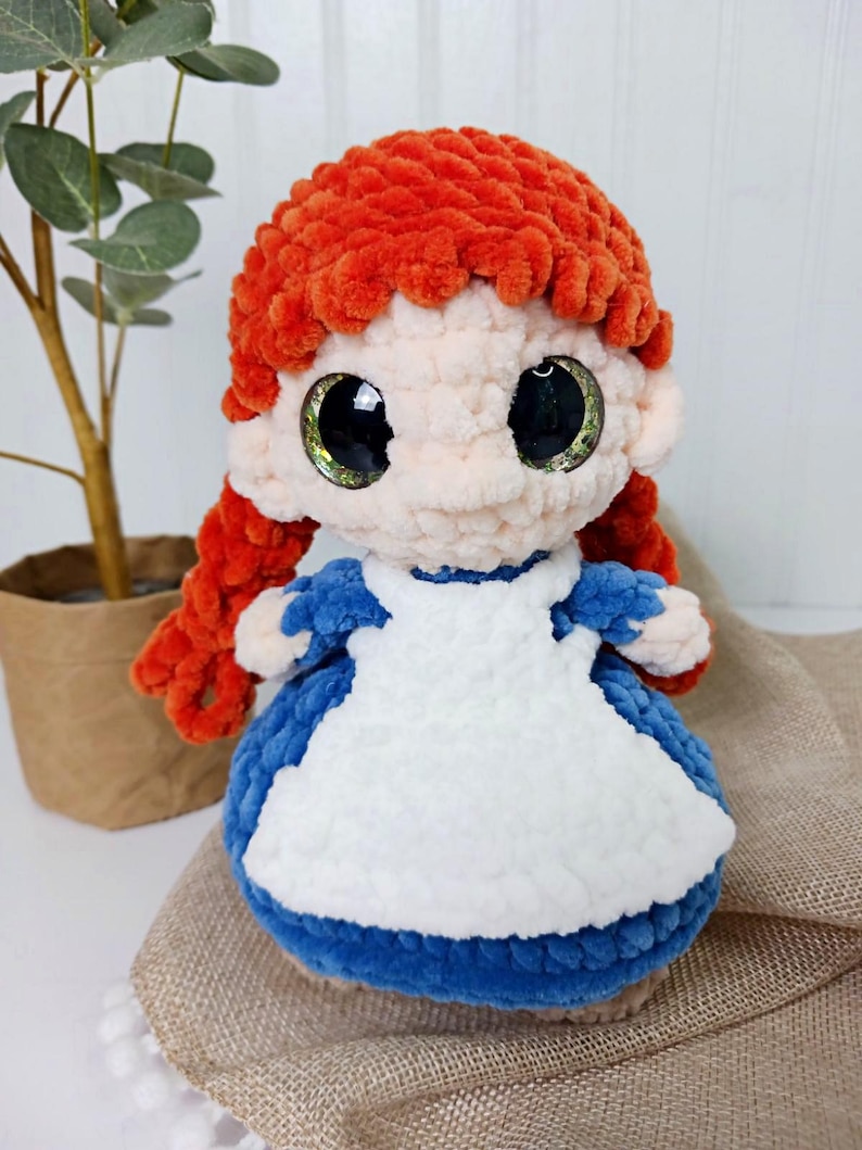 May include: A handmade plush doll with bright orange hair styled in a bun and pigtails. The doll has large, expressive eyes and is wearing a blue dress with a white apron. The doll is made of soft, textured yarn.