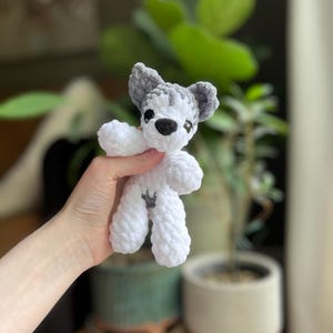 May include: A handmade crochet dog toy in white and gray. The toy has a small black nose and eyes, with gray ears and a gray patch on its back. The dog is made of soft, textured yarn.