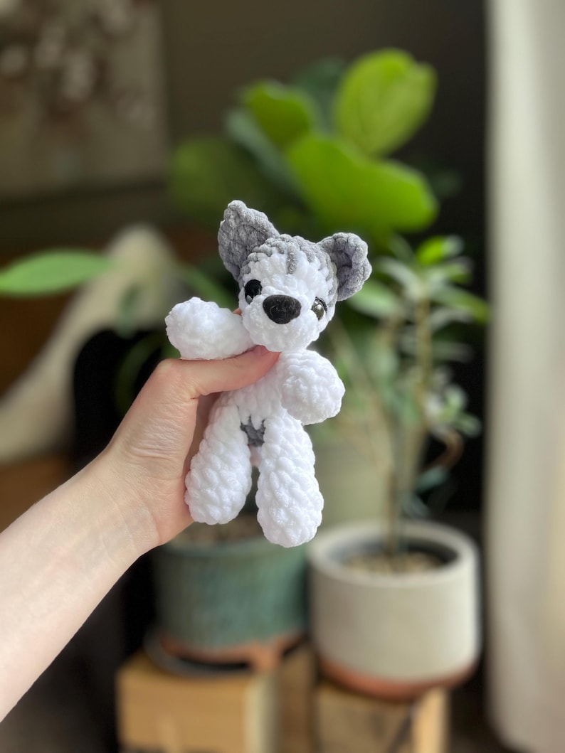 May include: A handmade crochet dog toy in white and gray. The toy has a small black nose and eyes, with gray ears and a gray patch on its back. The dog is made of soft, textured yarn.