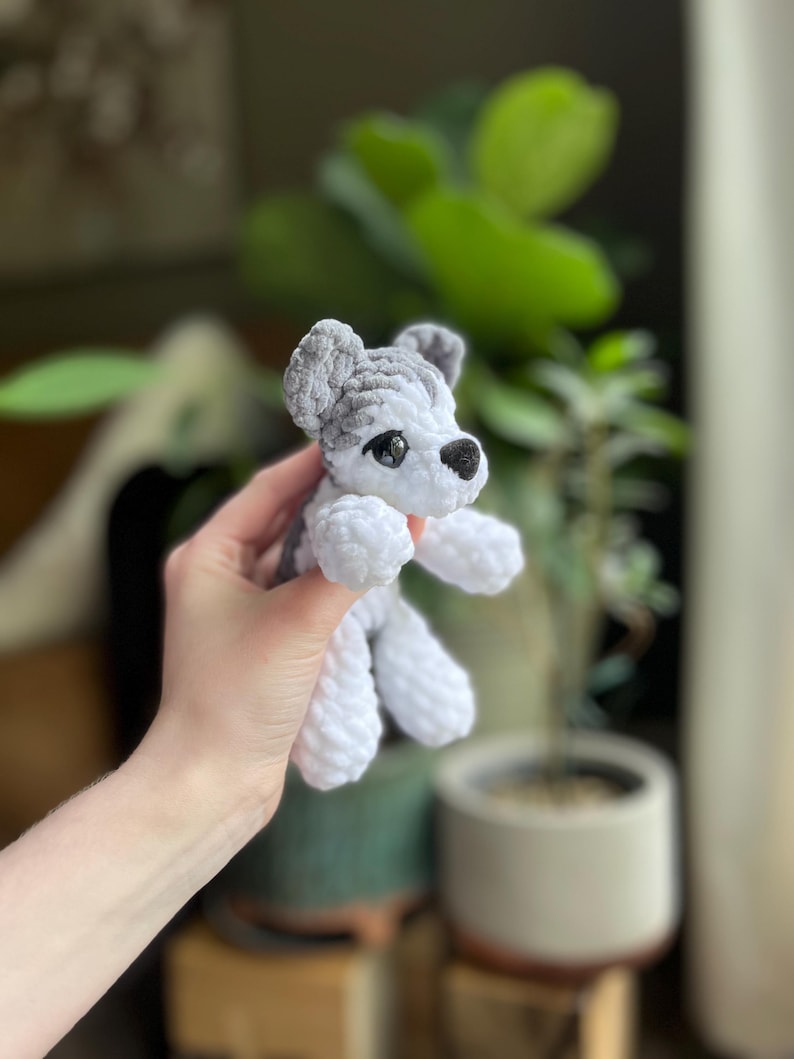 May include: A handmade crochet dog toy in white and gray. The dog has black eyes and a black nose. The toy is held in a person's hand. The background is blurred, with green plants and a neutral-colored pot.