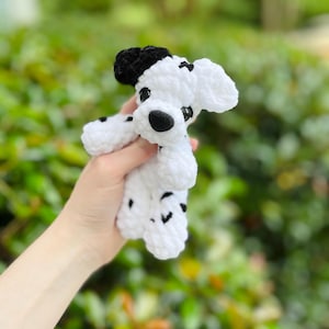 以下が含まれることがあります： ダルメシアン犬の形をした手編みの小さなかぎ針編みのぬいぐるみ。柔らかい白い毛糸に黒い斑点があり、片方の耳は黒、もう片方は白で、黒い鼻と暗く光沢のあるプラスチック製の目で作られています。