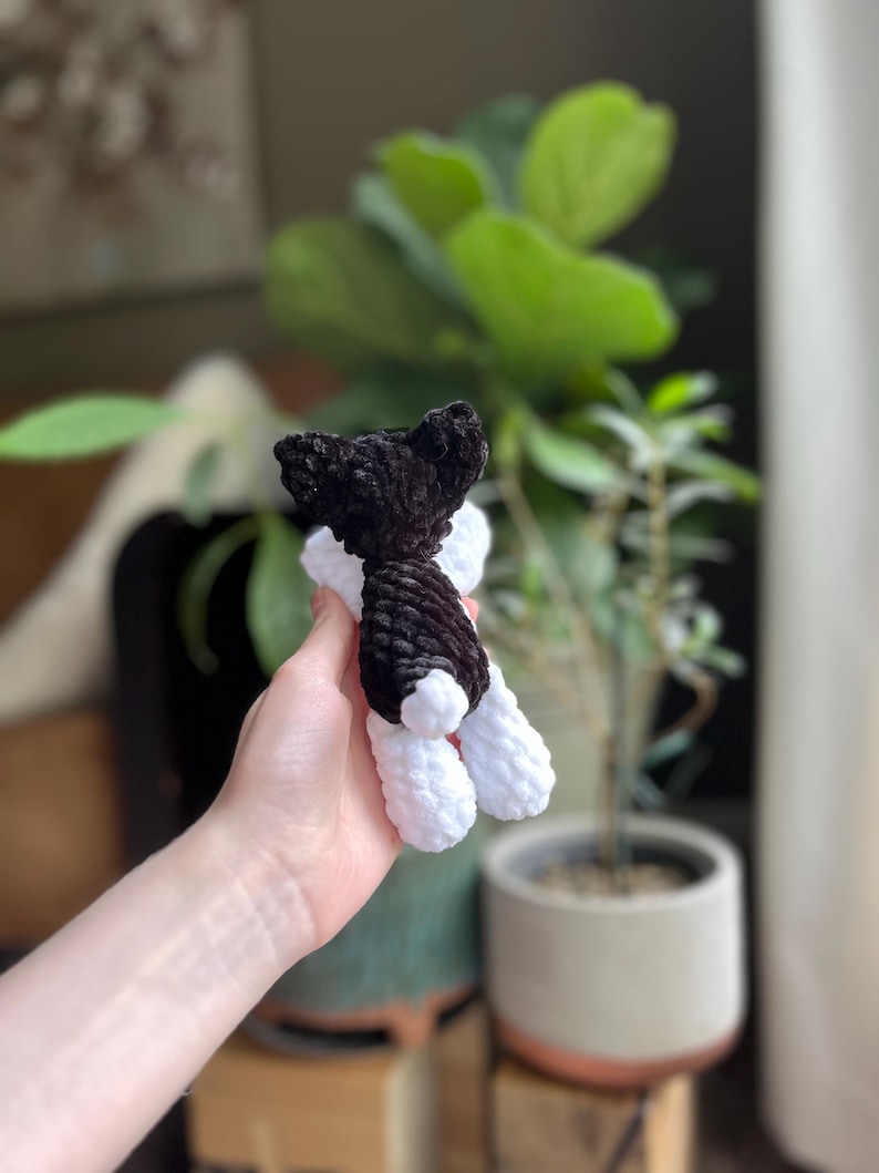 May include: A handmade crochet dog toy in black and white. The dog has a black head and body with white legs and feet. The toy is held in a person's hand, with a blurred background of plants.