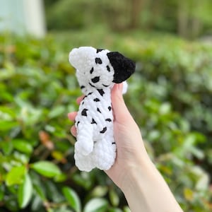 以下が含まれることがあります： ダルメシアン犬の形をした、手編みの小さなかぎ針編みのぬいぐるみ。白い毛糸で作られており、体全体に黒い斑点があり、片方の耳は無地の黒色で、かぎ針編み特有の柔らかく質感のある表面が特徴です。