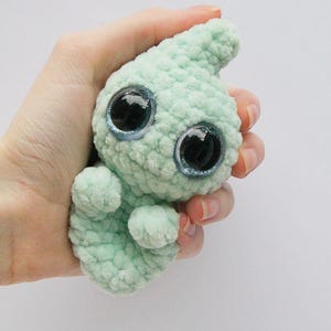 Puede incluir: Un peque&ntilde;o juguete de peluche de color verde claro tejido a crochet con ojos grandes y brillantes. El juguete tiene forma de l&aacute;grima y una superficie texturizada.