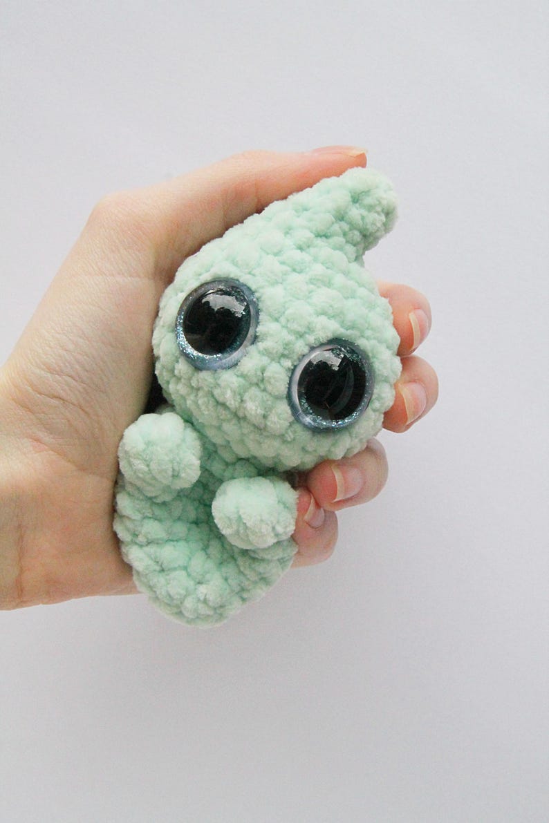 Puede incluir: Un peque&ntilde;o juguete de peluche de color verde claro tejido a crochet con ojos grandes y brillantes. El juguete tiene forma de l&aacute;grima y una superficie texturizada.