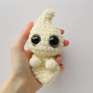 Puede incluir: Un fantasma blanco de crochet con grandes ojos negros y un borde dorado. El fantasma est&aacute; levantando las manos en un gesto amistoso.