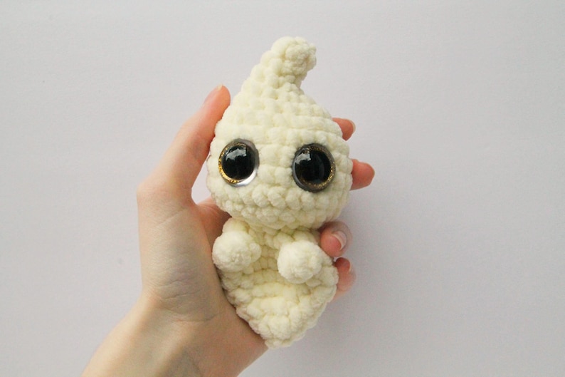 Puede incluir: Un fantasma blanco de crochet con grandes ojos negros y un borde dorado. El fantasma est&aacute; levantando las manos en un gesto amistoso.