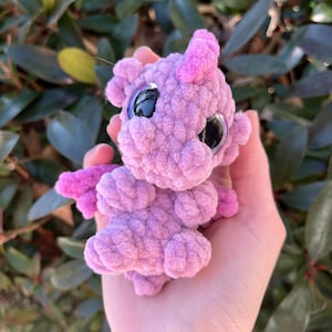May include: A small, handmade, lavender-colored crochet dragon with large, black, reflective eyes. The dragon has small pink horns and wings. The plush toy is held in a hand, showcasing its size and texture. The dragon is a whimsical, handcrafted item.