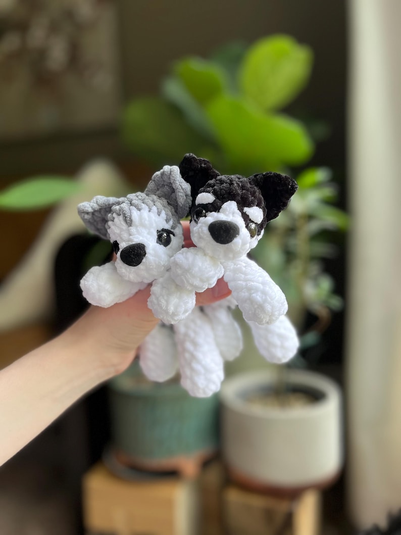 May include: Two handmade crochet dog toys. One dog is gray and white, the other is black and white. Both have black eyes and noses. The toys are held in front of a blurred background of plants and a pot.