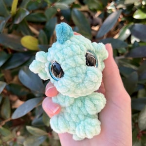 May include: A small, crocheted creature in shades of light blue and turquoise. It has large, reflective eyes and a small, upright fin on its head. The plush toy is held in a hand against a backdrop of green foliage.
