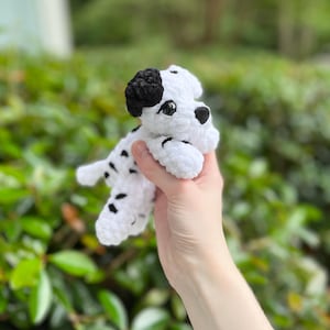 以下が含まれることがあります： ダルメシアン犬の形をした手編みの小さなかぎ針編みのぬいぐるみ。白い毛糸に黒い斑点があり、片方の黒い耳、黒い鼻、暗い丸いプラスチック製の目が特徴です。質感は柔らかく、ふわふわとしています。