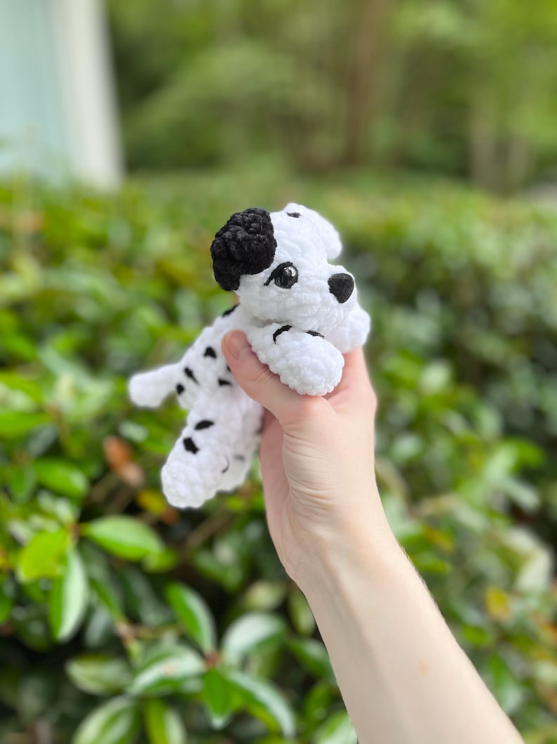 以下が含まれることがあります： ダルメシアン犬の形をした手編みの小さなかぎ針編みのぬいぐるみ。白い毛糸に黒い斑点があり、片方の黒い耳、黒い鼻、暗い丸いプラスチック製の目が特徴です。質感は柔らかく、ふわふわとしています。
