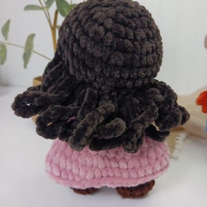May include: A handmade crochet doll with dark brown hair and a pink dress. The doll's hair is styled in a curly, textured design. The dress is a soft pink color, and the doll has brown shoes. The doll is a handmade item.