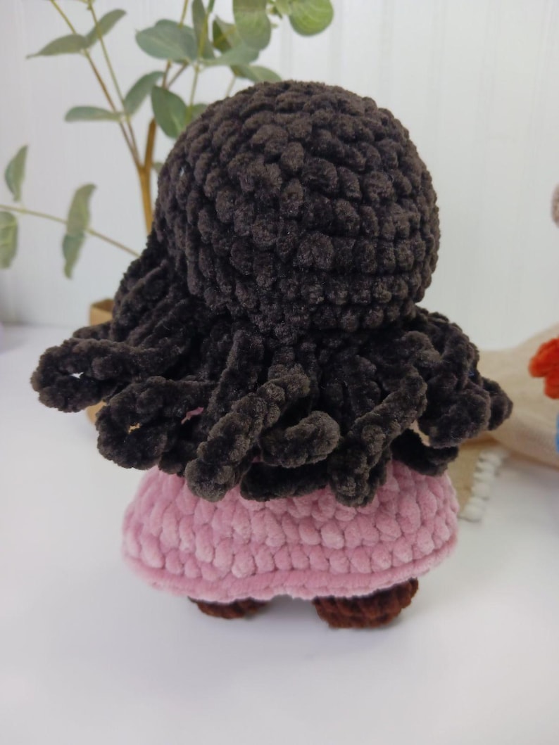 May include: A handmade crochet doll with dark brown hair and a pink dress. The doll's hair is styled in a curly, textured design. The dress is a soft pink color, and the doll has brown shoes. The doll is a handmade item.