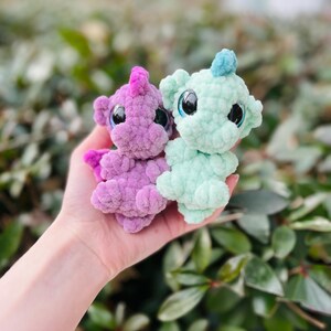 May include: Two handmade crochet dragon plushies. One is lavender with pink accents, the other is mint green with a blue accent. Both have large, black-pupiled eyes. The plushies are held in a hand against a blurred green background.