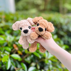 以下が含まれることがあります： 柔らかい質感の糸で作られた2匹の小さな犬のぬいぐるみ。1匹はライトベージュ、もう1匹はミディアムブラウンです。どちらも丸い黒いプラスチックの目、小さく丸い黒いフェルトの鼻、垂れ耳、そしてシンプルなステッチの口が特徴です。