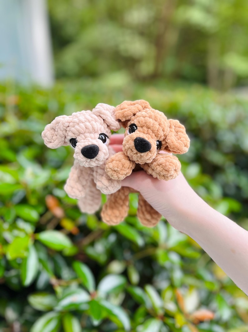 以下が含まれることがあります： 柔らかい質感の糸で作られた2匹の小さな犬のぬいぐるみ。1匹はライトベージュ、もう1匹はミディアムブラウンです。どちらも丸い黒いプラスチックの目、小さく丸い黒いフェルトの鼻、垂れ耳、そしてシンプルなステッチの口が特徴です。