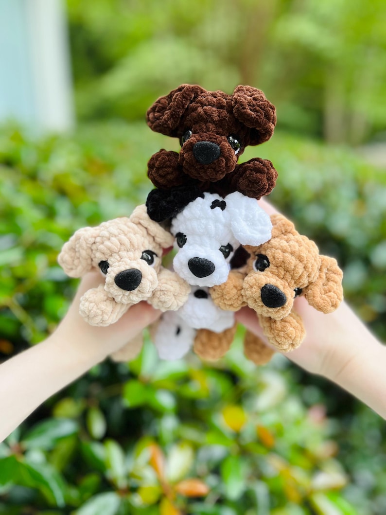 以下が含まれることがあります： 柔らかい質感の糸で編まれた、4匹の小さな犬のぬいぐるみ。ダークブラウン、ライトベージュ、黒いアクセントのある白、タンカラーの犬たちで、それぞれ黒いプラスチックの目と黒い刺繍の鼻が特徴です。