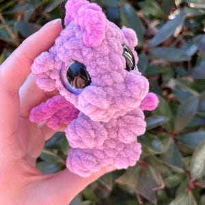 May include: A handmade, plush dragon toy in shades of pink and purple. The dragon has large, reflective eyes, small wings, and a textured, crocheted body. The toy is held in a hand against a backdrop of green foliage.