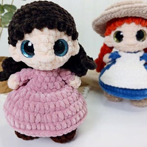 May include: Two handmade crochet dolls. One doll has black hair and a pink dress, the other has red hair, a hat, and a blue and white dress. Both dolls have large, shiny eyes.