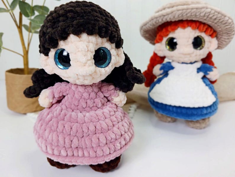 May include: Two handmade crochet dolls. One doll has black hair and a pink dress, the other has red hair, a hat, and a blue and white dress. Both dolls have large, shiny eyes.