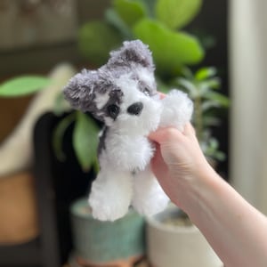 May include: A small, plush toy dog with a black nose and black and gray fur. The toy dog has white fur on its paws and belly. The toy is held in front of a blurred background of green plants and pots.