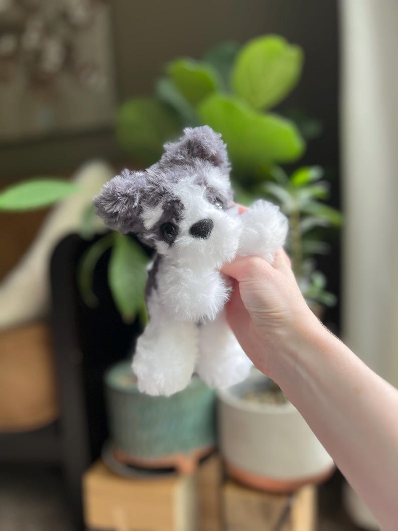 May include: A small, plush toy dog with a black nose and black and gray fur. The toy dog has white fur on its paws and belly. The toy is held in front of a blurred background of green plants and pots.