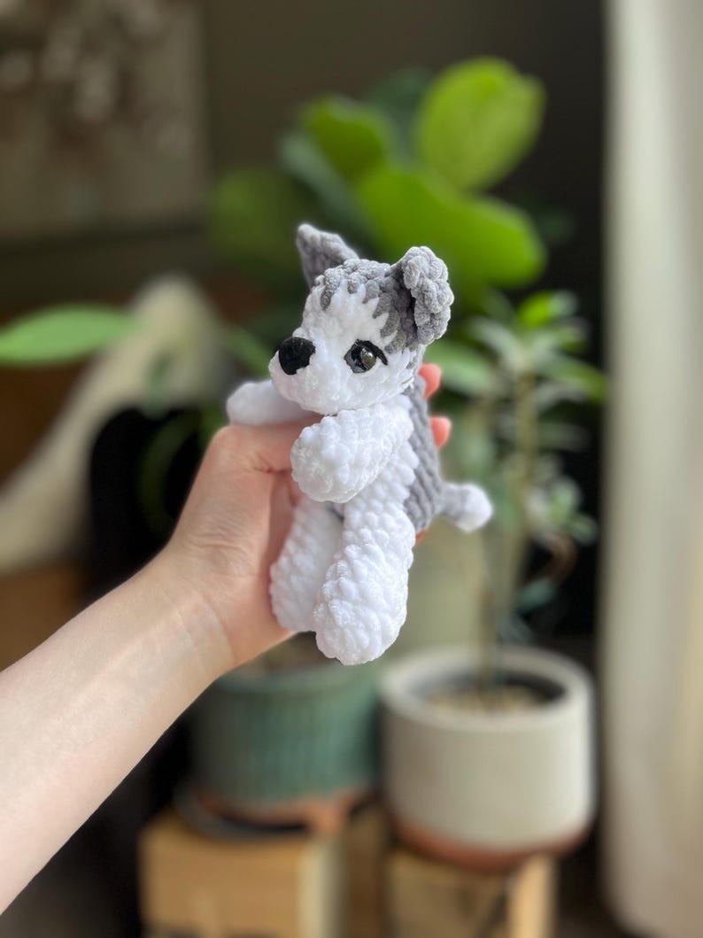 May include: A small, handmade crochet dog toy in shades of white and gray. The plush toy features a detailed head with black eyes and a small black nose. The dog's body is white, and the ears and a patch on the back are gray.