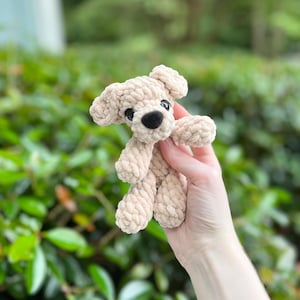 以下が含まれることがあります： 厚みのある質感の糸で編まれた、ベージュ色の小さなかぎ針編みの犬のぬいぐるみ。このぬいぐるみには、垂れ下がった耳が2つ、黒い鼻が付いた丸みを帯びた鼻先、そして2つの小さくて暗い丸い目があります。